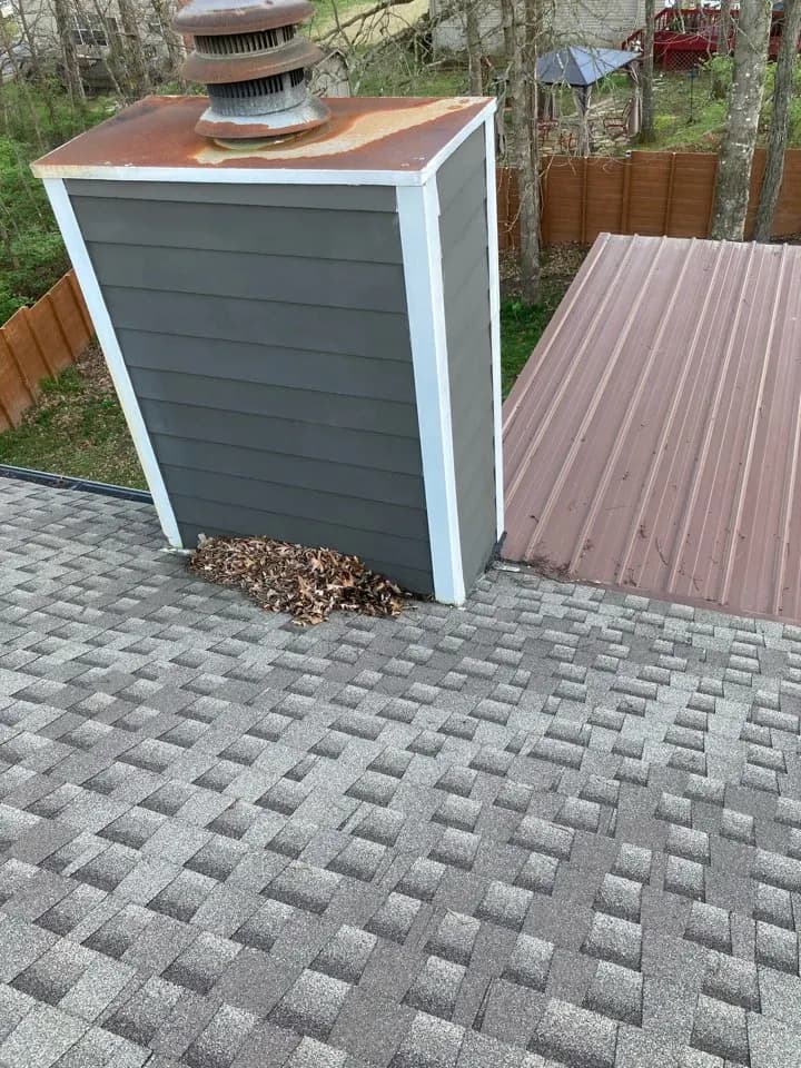 Shingle roof section beside a sided chimney chase with leaves collected at its base, during a leaking chimney repair in Nashville