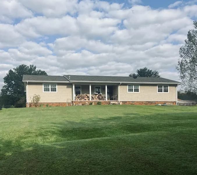 Single-story ranch style home with beige siding, brick foundation accents, and a covered front porch, set on a large grassy lawn under a partly cloudy sky
