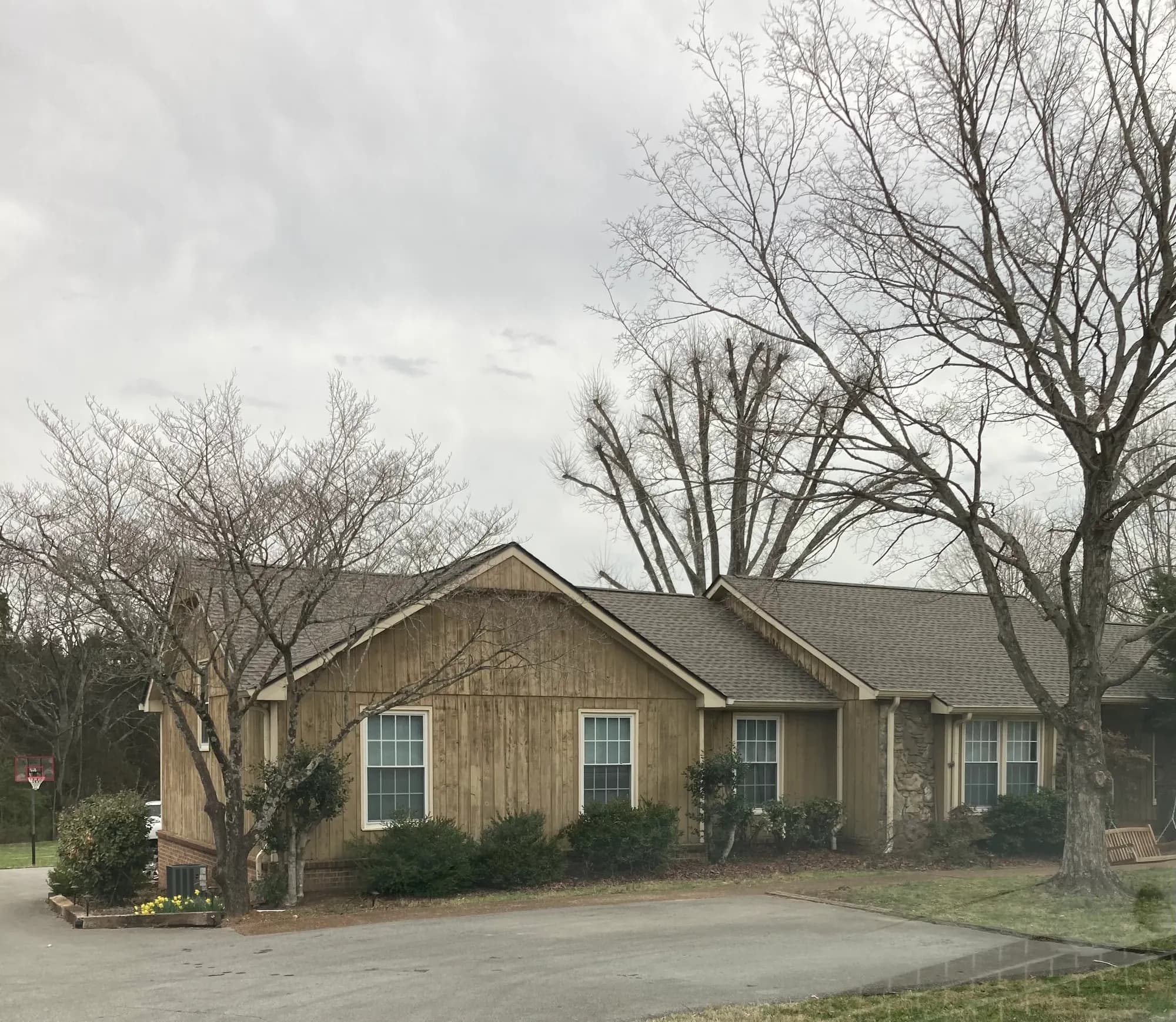 A single-story house with wood board-and-batten siding and a newly installed asphalt shingle roof, surrounded by bare trees