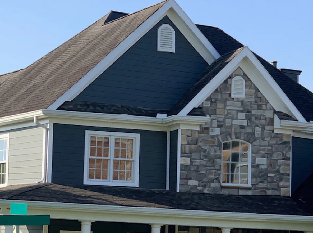 Closer view of the roof: a large two-story home with dark gray roofing shingles, stone accents, and a covered front porch, set under a clear blue sky