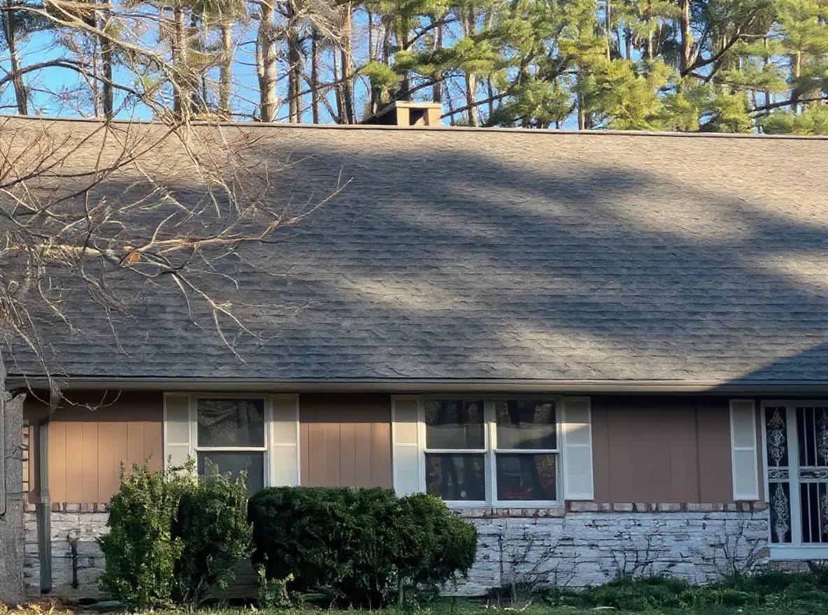 A single-story brick and wood-sided ranch home with a composition shingle roof, surrounded by mature trees on a large grassy lot